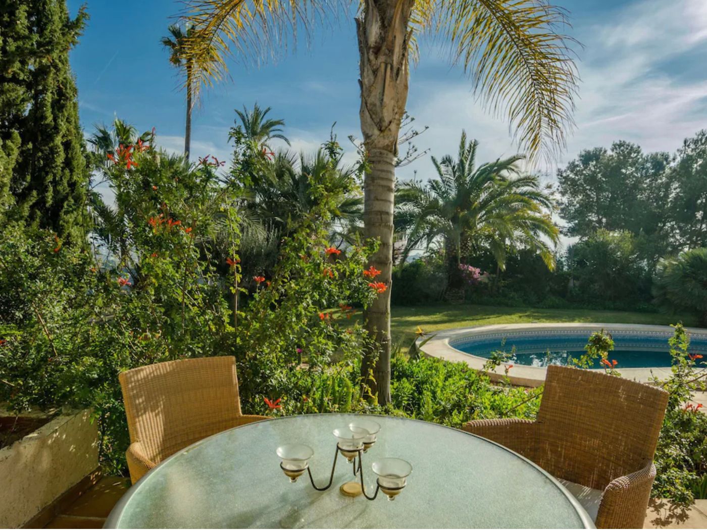 Outdoor terrace featuring a glass-topped table and wicker seating, overlooking a circular swimming pool and lush Mediterranean gardens with mature palm trees.