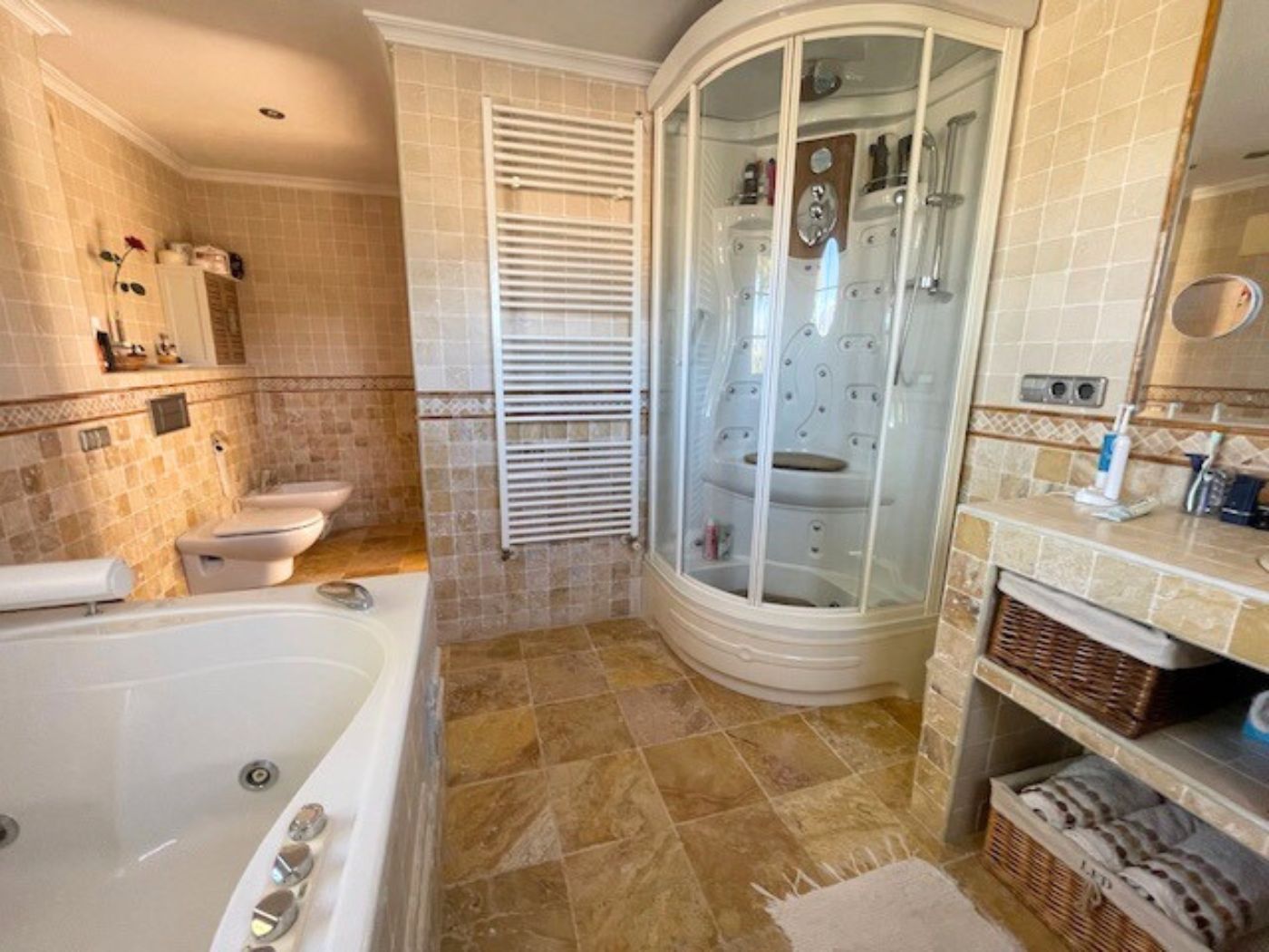Spacious bathroom featuring a corner hydromassage shower cabin, a large bathtub, and natural stone tiling throughout. Includes a heated towel rail and wicker storage.
