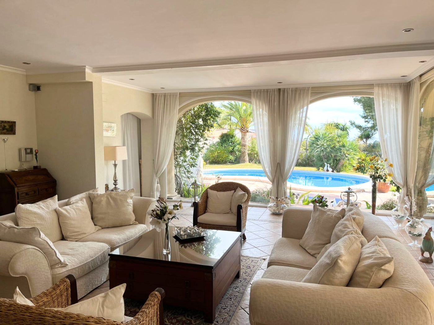 Spacious living room featuring cream-toned sofas and wicker seating, with large arched windows offering direct views of the private pool and Mediterranean garden.