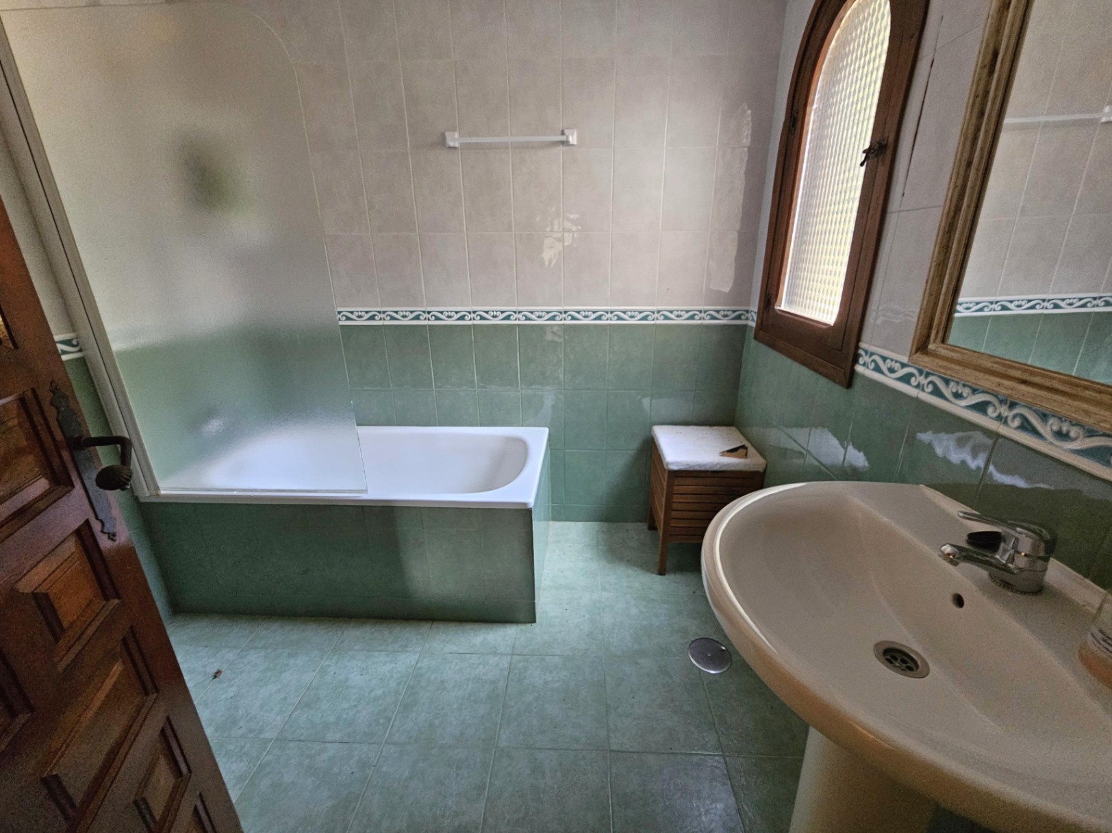 Bathroom featuring a built-in bathtub with a frosted glass screen, green ceramic tiling with a decorative border, and a wooden arched window.