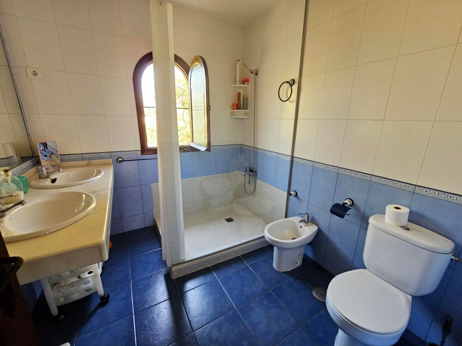 Bathroom featuring a double marble vanity, walk-in mosaic shower, bidet, and toilet. Includes a wooden arched window and blue and white tiled walls.