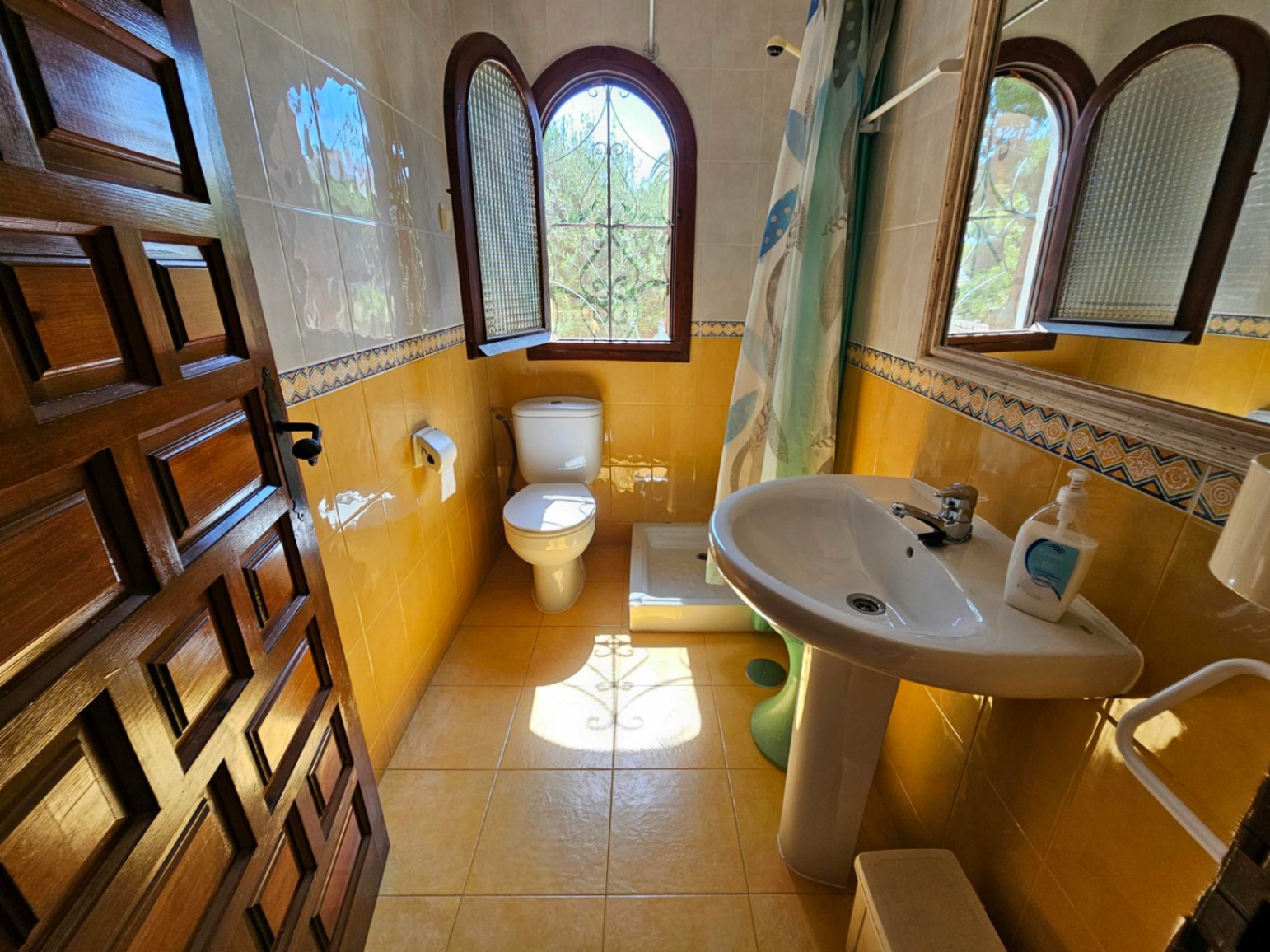 Bathroom featuring yellow wall tiling with decorative borders, a white pedestal sink, and an arched wooden window with wrought iron.