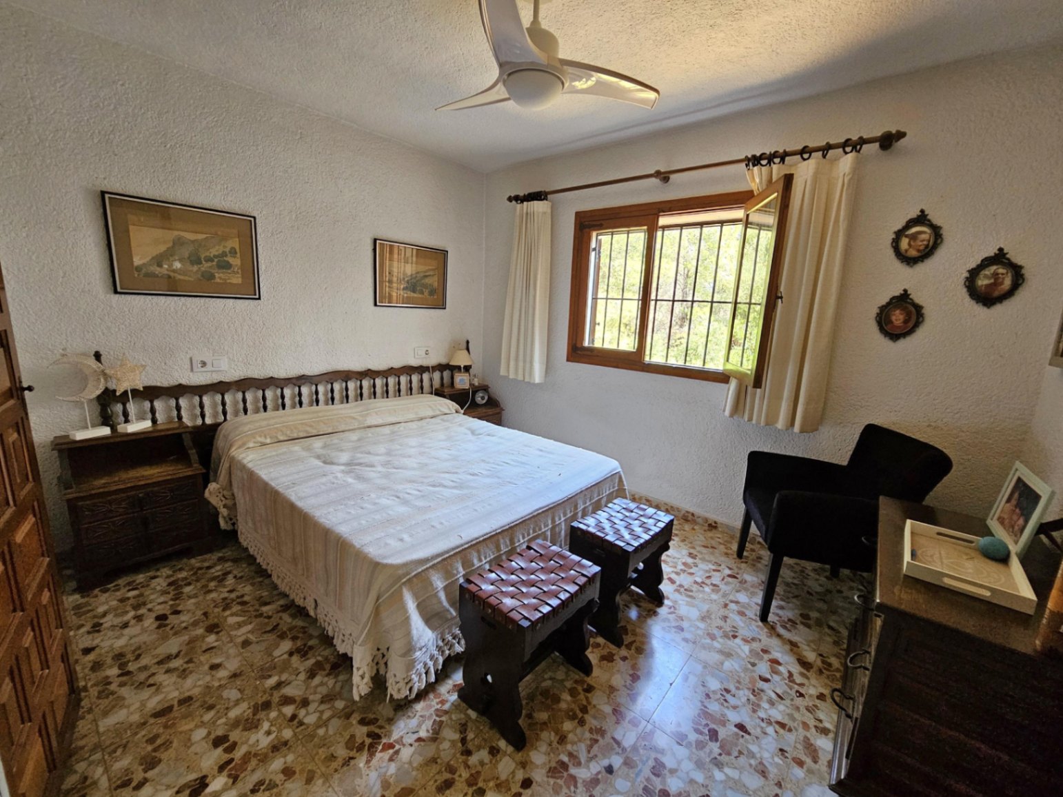 Bedroom featuring traditional terrazzo flooring, a wooden spindle headboard, and dual leather-topped stools. Natural light enters through a wooden-framed window.