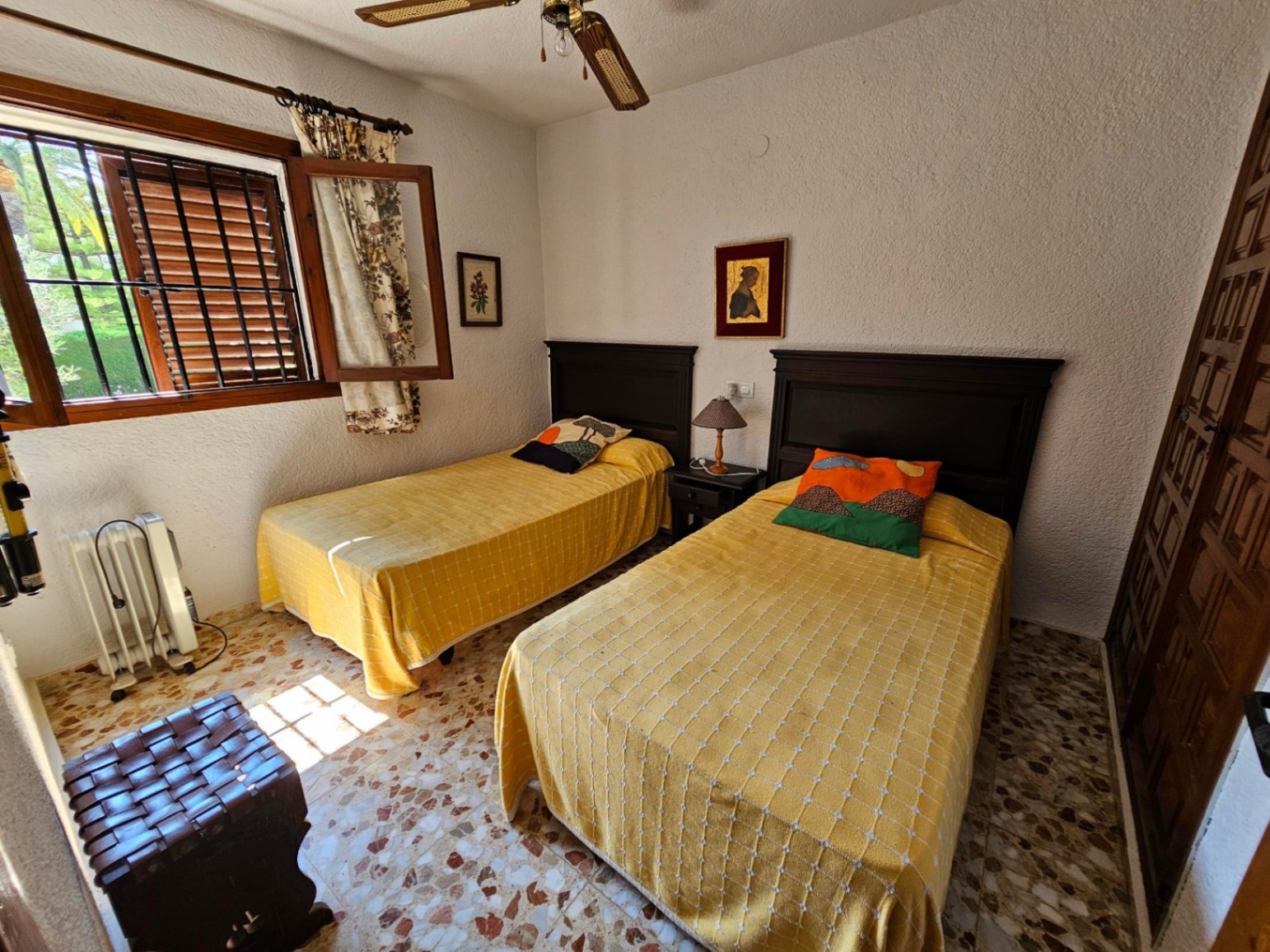 Bedroom featuring twin beds with dark wood headboards, yellow linens, and classic terrazzo flooring. Includes a ceiling fan and natural light from a barred window.