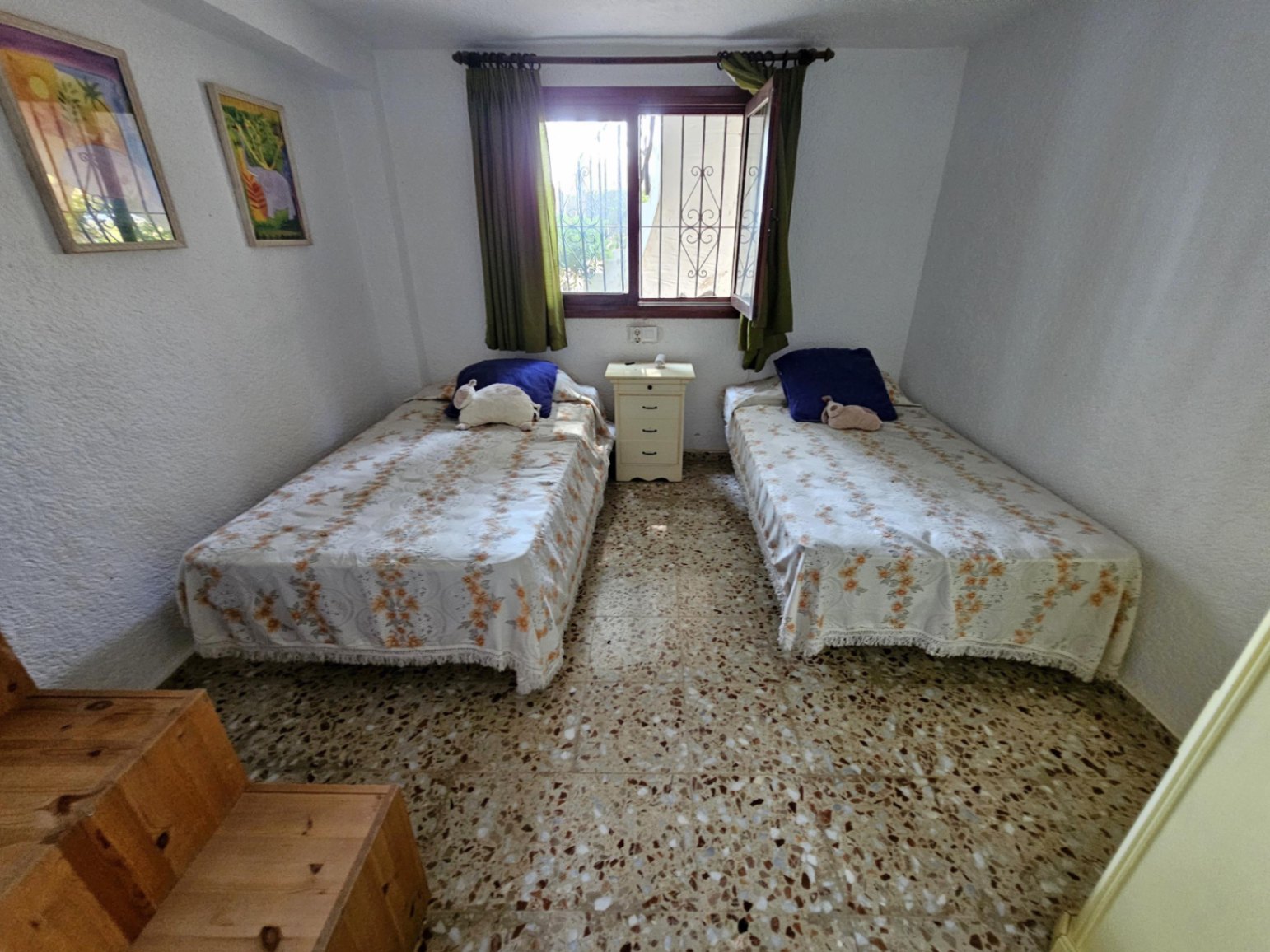 Bedroom featuring twin beds with floral covers, terrazzo flooring, and a central nightstand. A window with security bars and green curtains allows natural light.