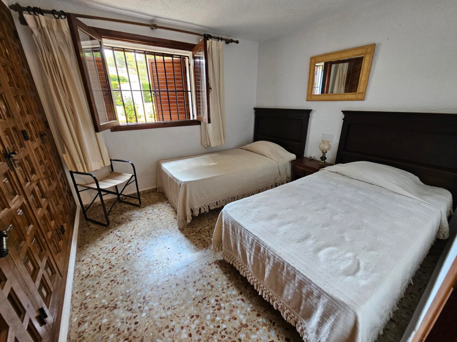 Bedroom with twin beds, dark wood headboards, and terrazzo flooring. Features a built-in wooden wardrobe and a window with security bars and shutters.