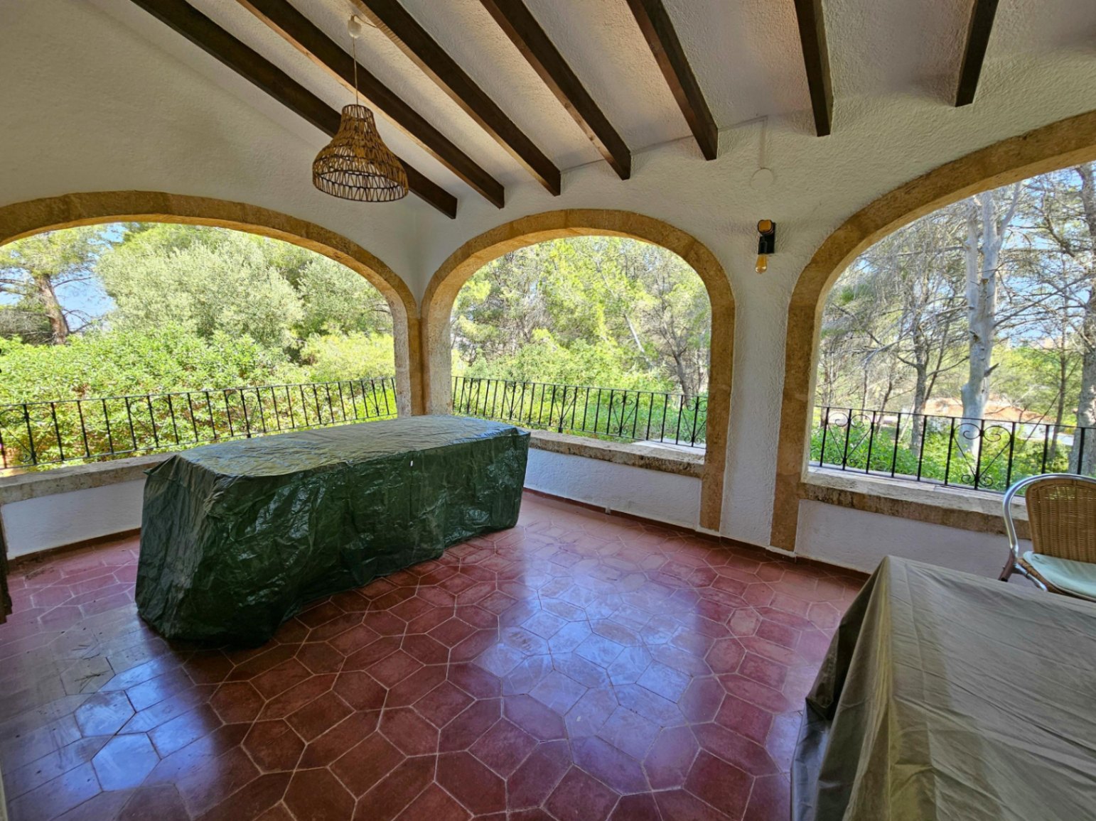Covered terrace featuring exposed wooden beams, red hexagonal terracotta flooring, and stone arches overlooking a lush wooded area.
