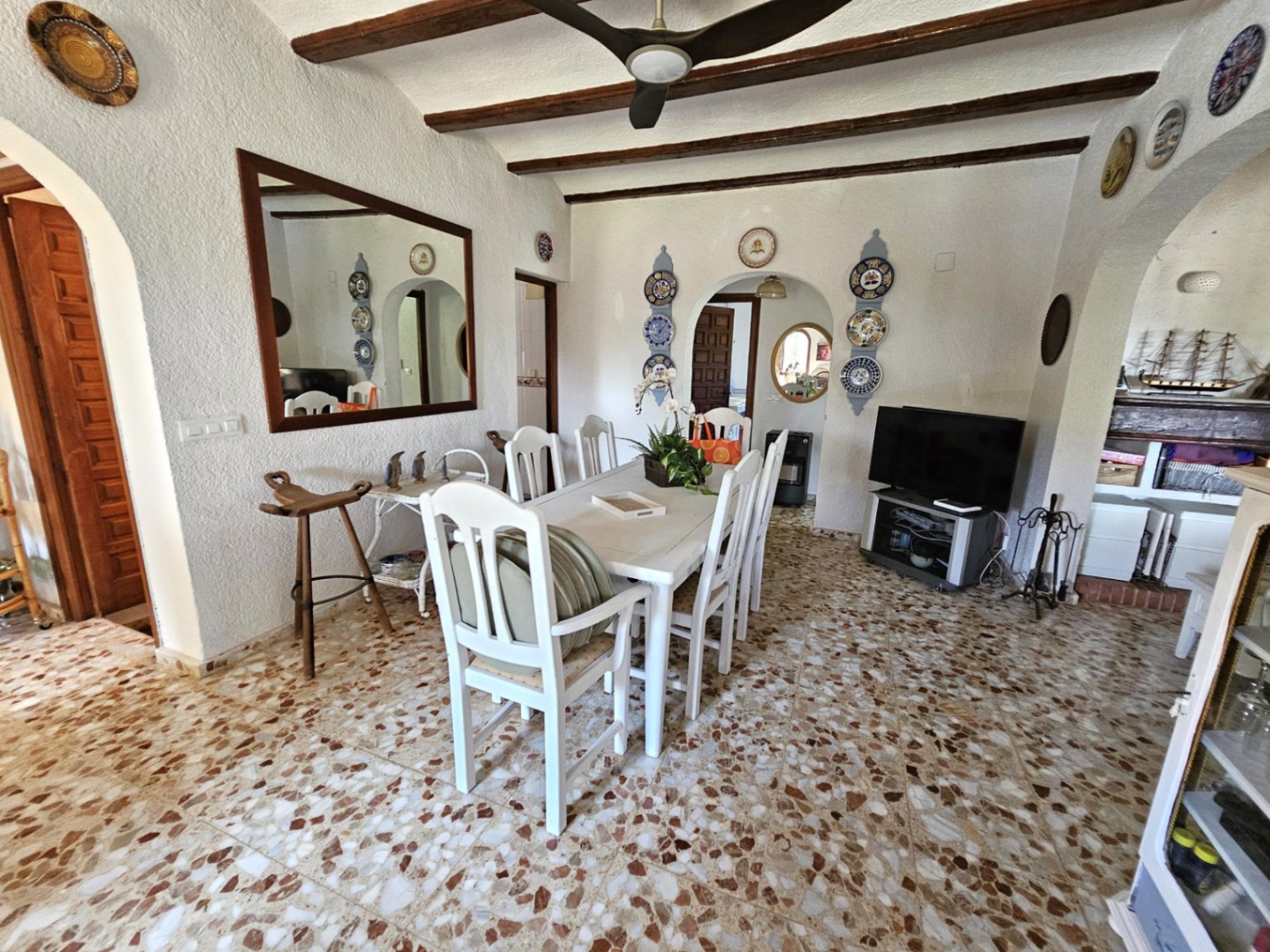 Dining area featuring a white wooden table set, exposed ceiling beams, and traditional terrazzo flooring. Decorative ceramic plates and arched doorways add rustic character.