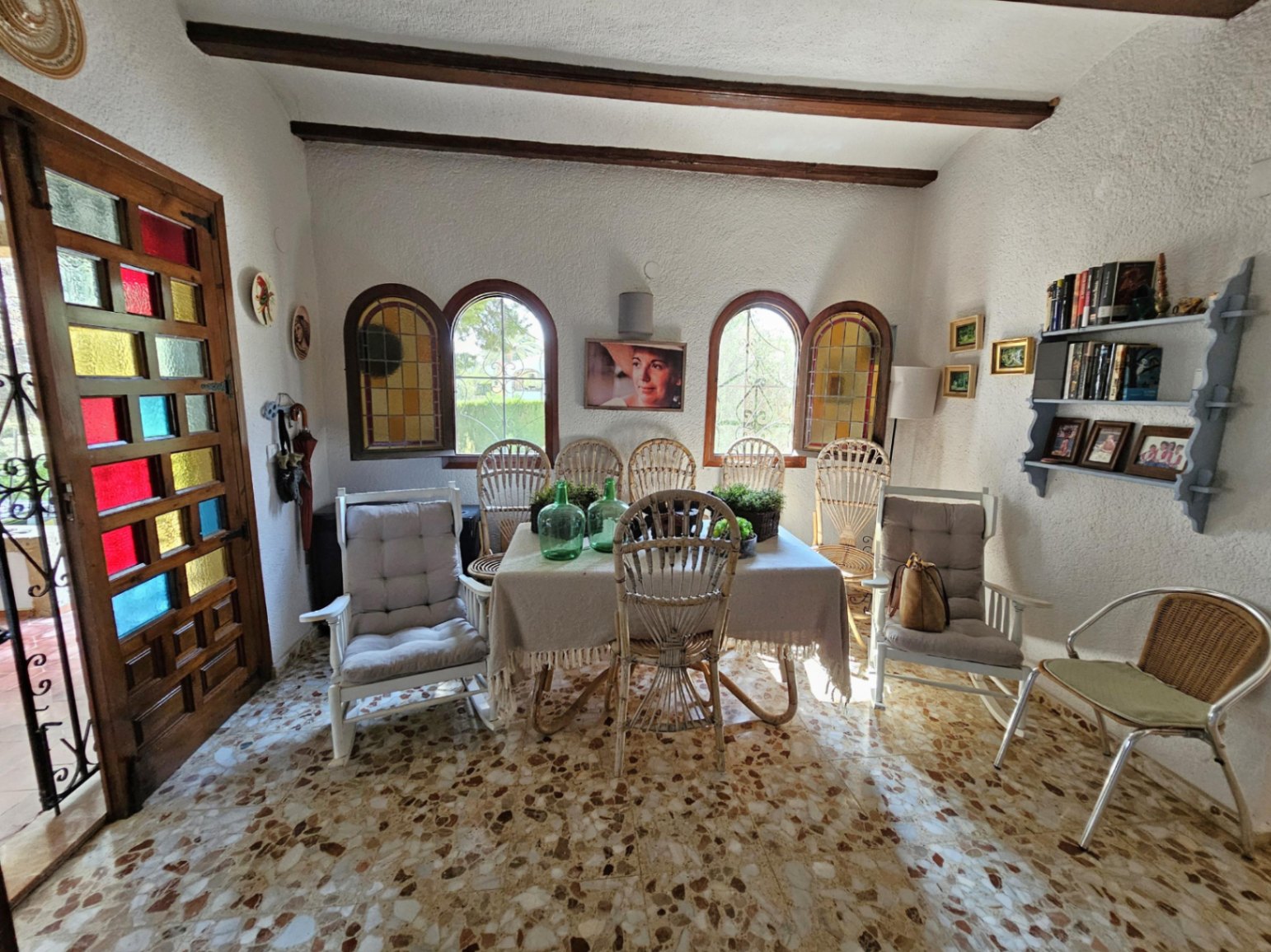 Dining area featuring terrazzo flooring, exposed wooden ceiling beams, and arched windows. Includes a wooden door with colored glass panes and rattan seating.