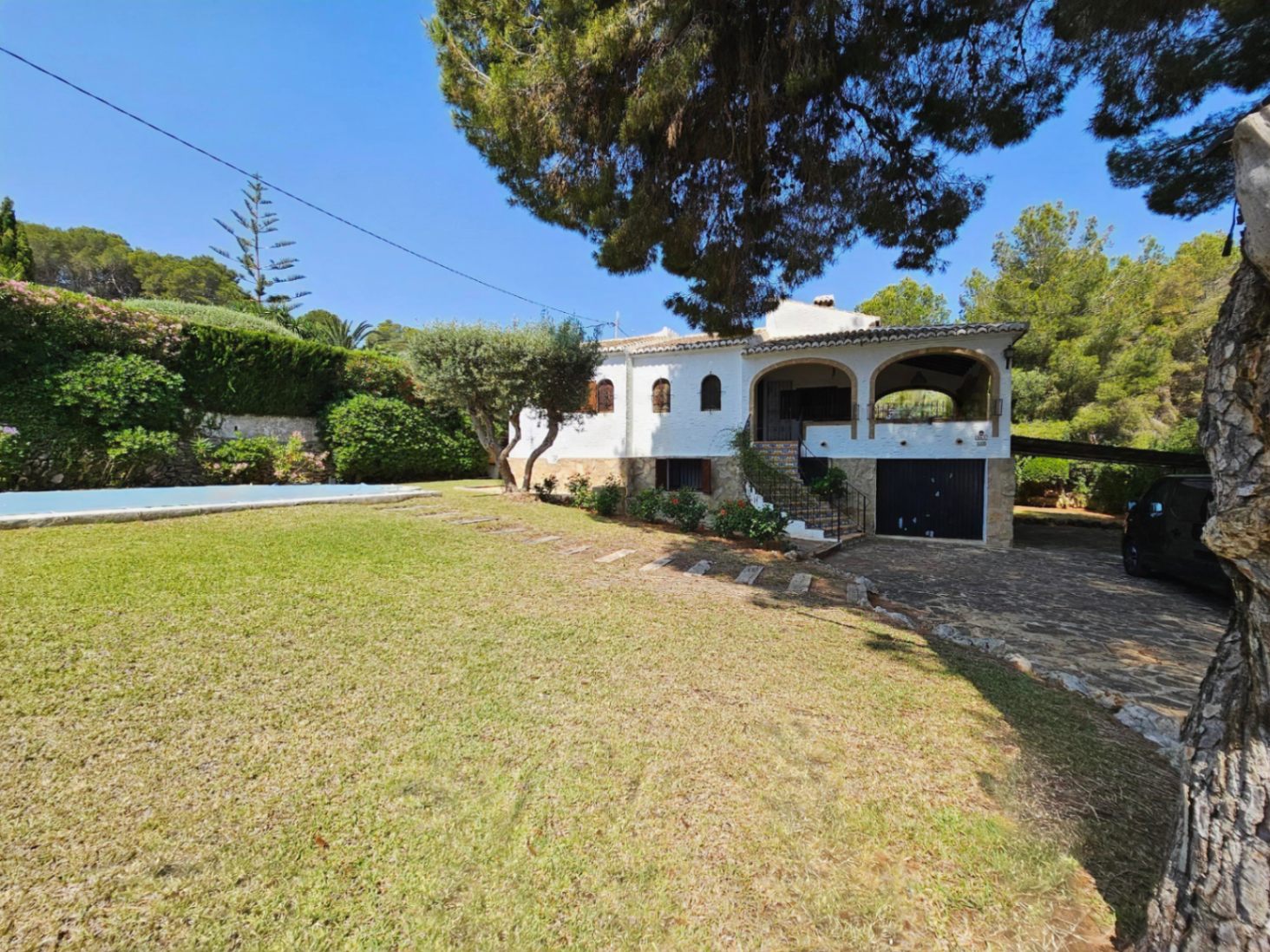 Exterior of a white Mediterranean villa featuring an arched porch, stone driveway, and manicured lawn adjacent to a swimming pool and mature pine trees.