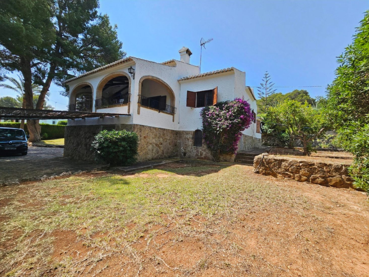 Exterior view of a Mediterranean villa featuring a stone-clad base, white facades, and a traditional arched terrace. The garden includes mature trees and bougainvillea.