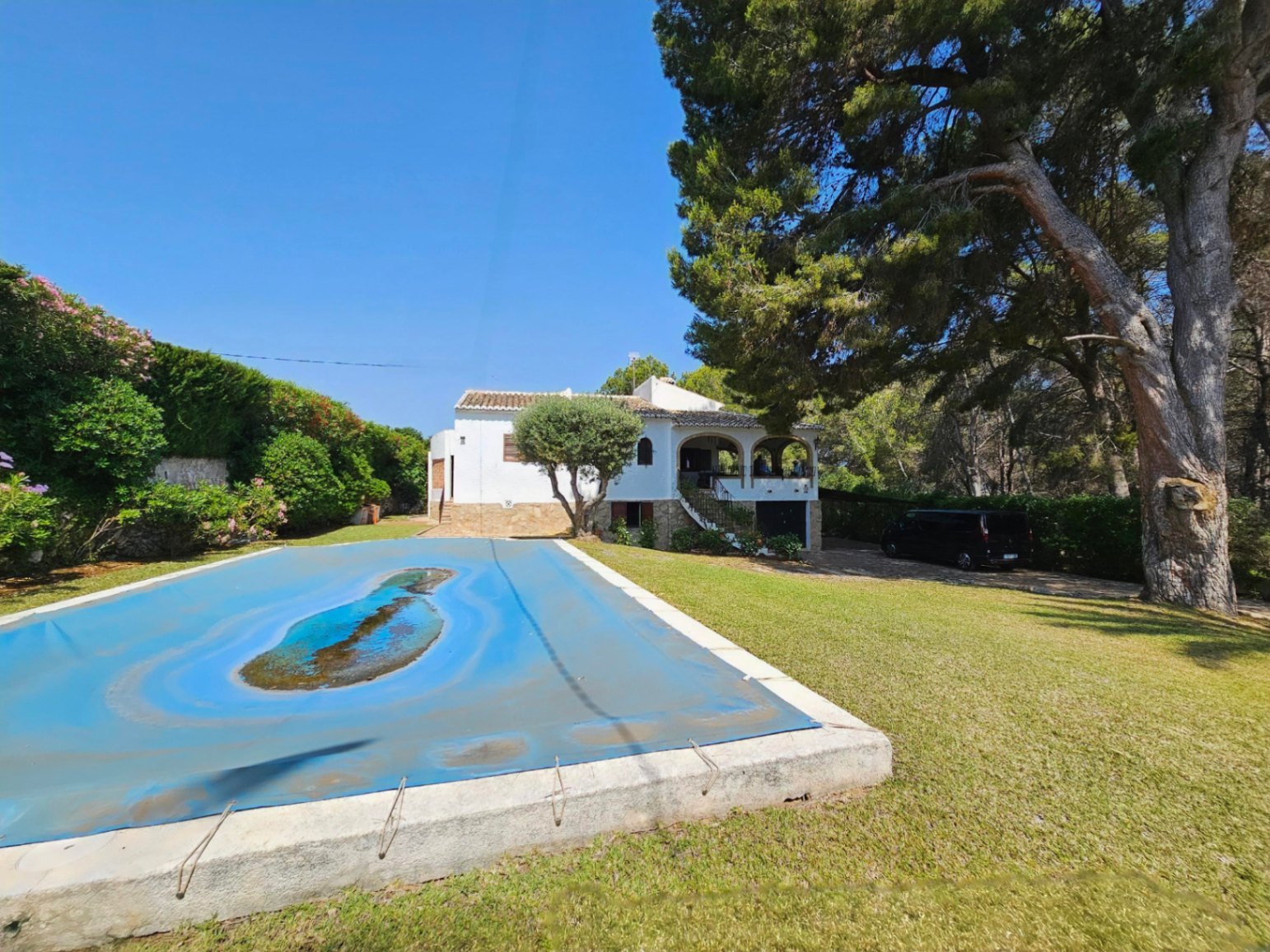 Exterior view of a white villa featuring an arched porch and stone foundation, set within a lawned garden with mature pine trees and a covered swimming pool.