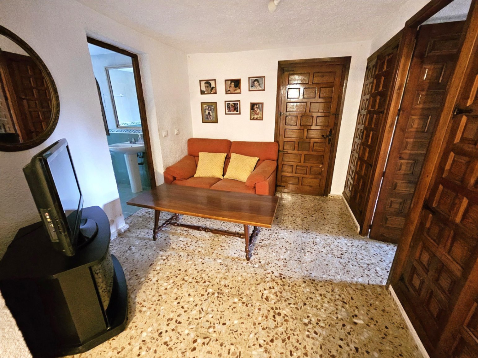 Living area featuring a terracotta sofa, carved wooden doors, and traditional terrazzo flooring. A doorway reveals a bathroom with a pedestal sink.