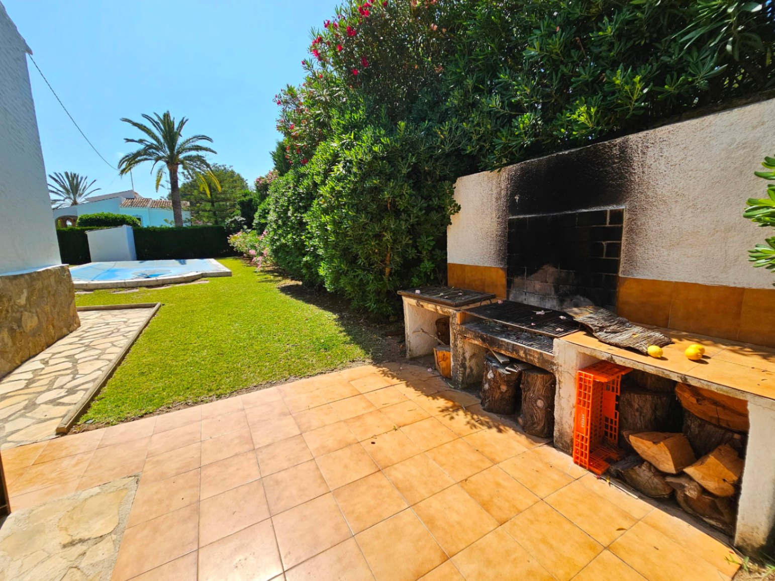 Outdoor barbecue station with terracotta tiling and wood storage, overlooking a manicured lawn, covered swimming pool, and Mediterranean palms.