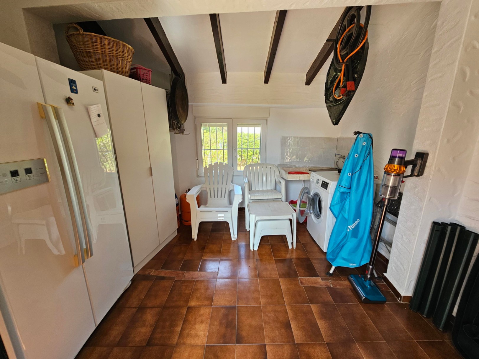 Practical utility room featuring terracotta tile flooring, exposed dark wood ceiling beams, a large refrigerator, laundry facilities, and integrated storage cabinets.