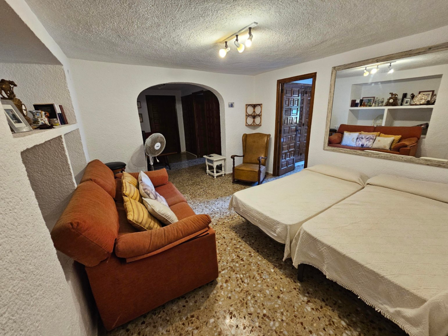 Spacious room featuring terrazzo flooring, two twin beds, an orange sofa, and built-in wall niches. Includes an arched entryway and a large framed mirror.