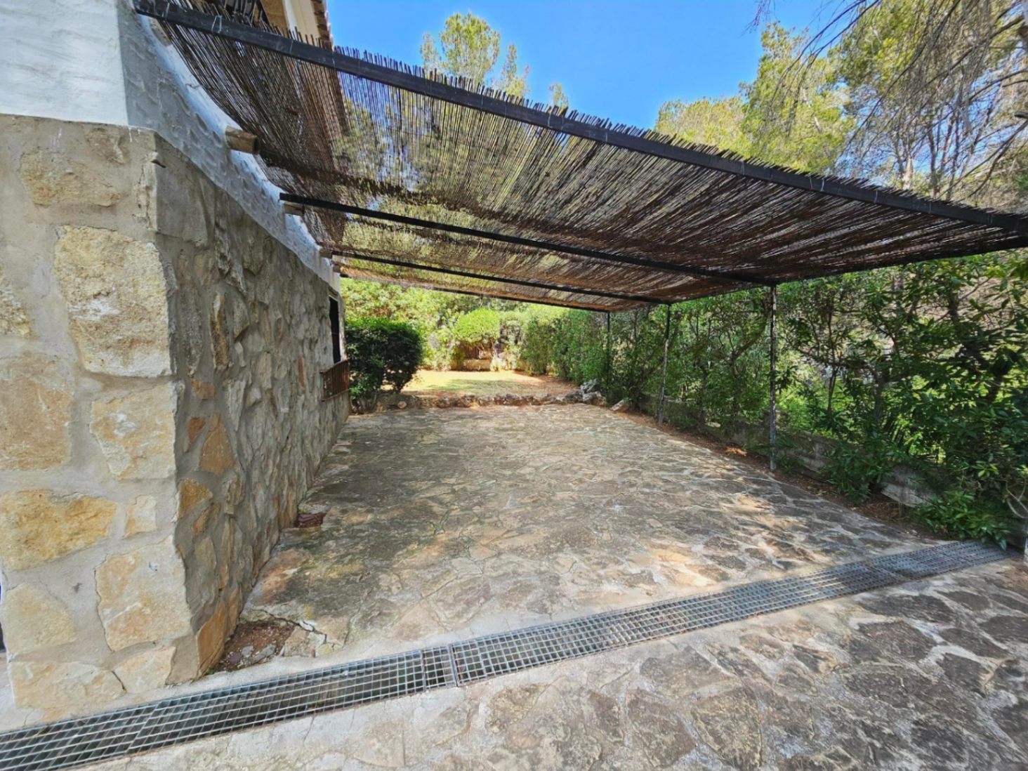 Terrace with irregular stone paving and a natural stone wall, featuring a shaded pergola with a reed roof overlooking the garden.