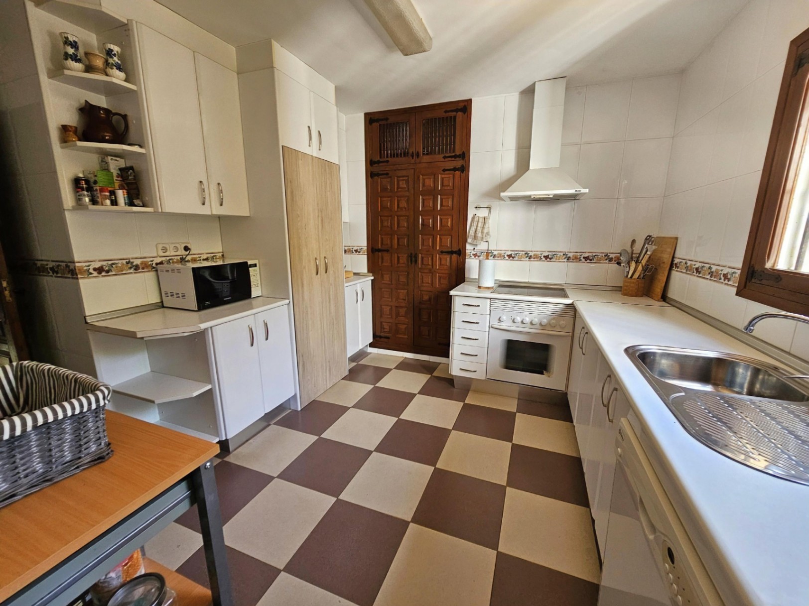 Traditional kitchen with checkered flooring, white cabinetry, and a carved dark wood door. Features a stainless steel sink and white appliances.