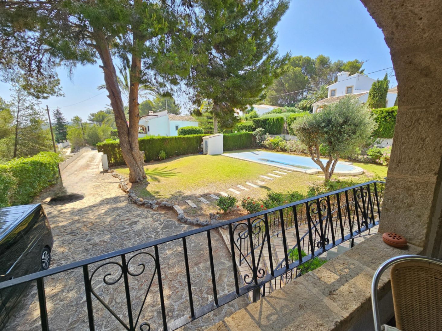 View from a stone terrace with wrought-iron railings overlooking a lawn, swimming pool, and stone-paved driveway surrounded by Mediterranean pines.