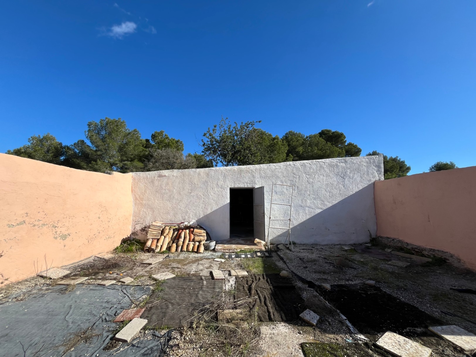 Enclosed courtyard featuring a white-washed outbuilding, traditional terracotta roof tiles, and peach-colored perimeter walls under a clear sky.