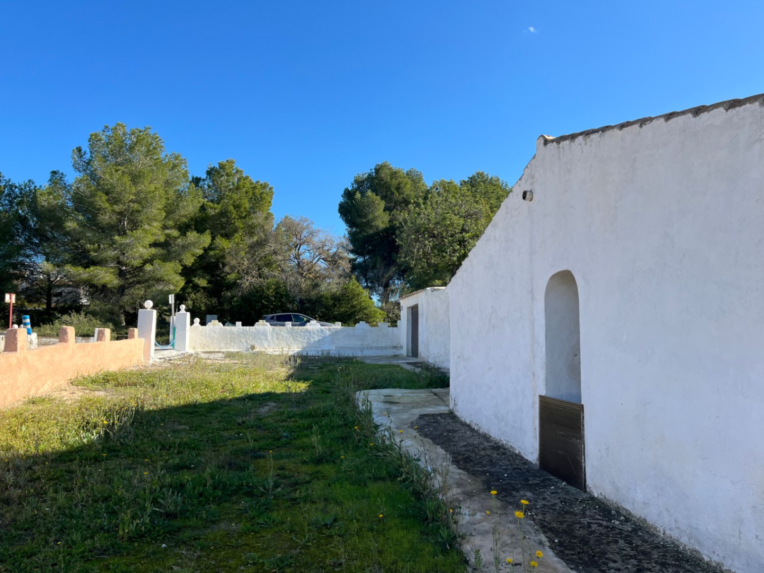 Exterior of a traditional white finca in Jávea, featuring an arched window niche and a natural garden area surrounded by mature pine trees.
