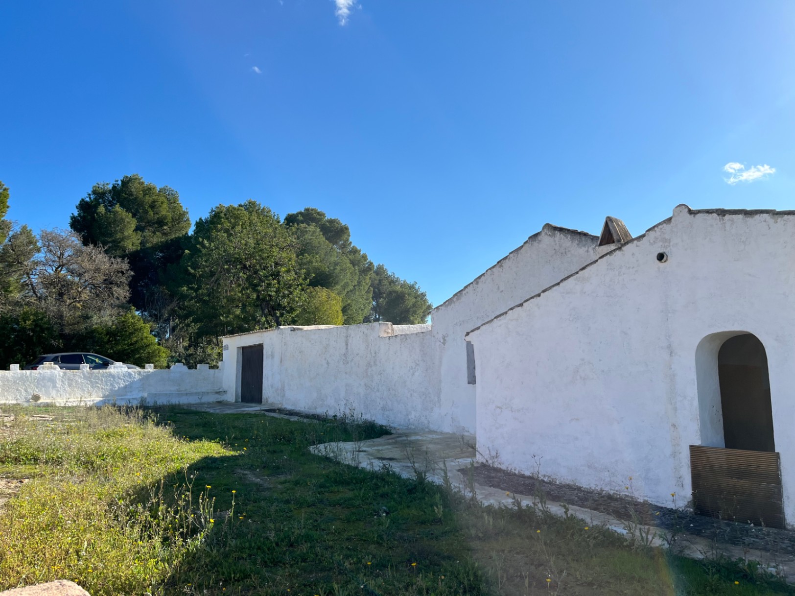 Exterior view of a traditional white-washed finca in Jávea, featuring an arched entrance and original stone walls surrounded by pine trees.