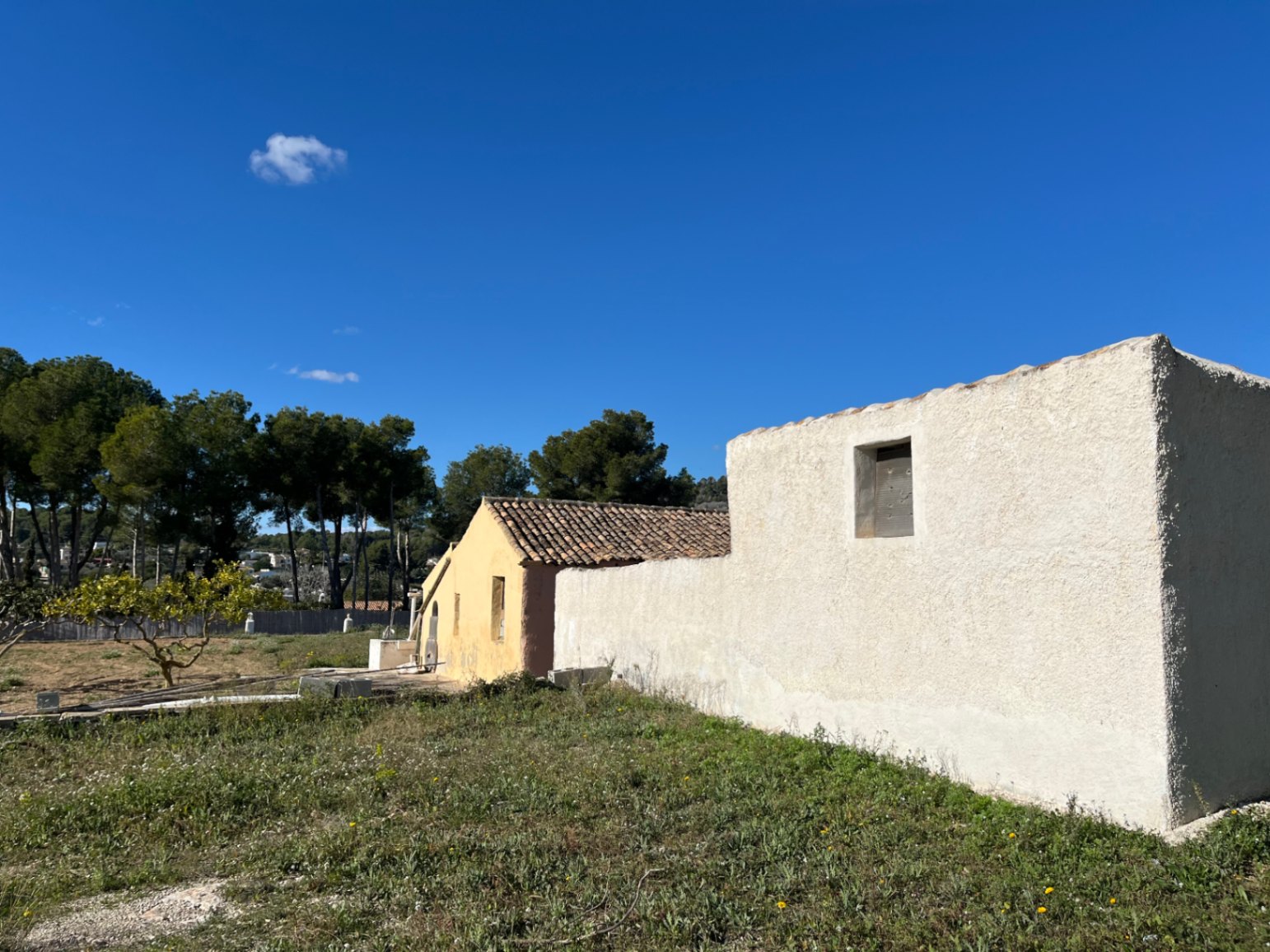 Exterior view of the traditional finca showing textured white and ochre walls, terracotta roof tiles, and surrounding natural plot under a clear sky.