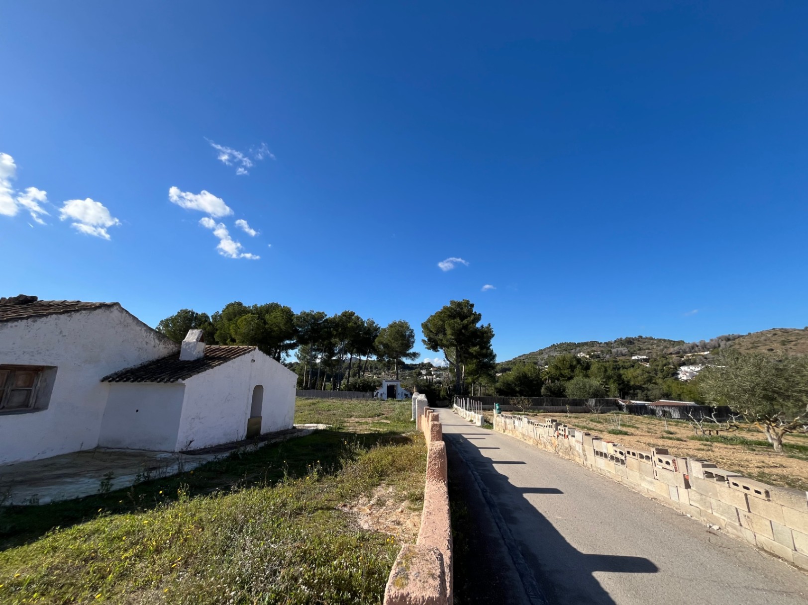 View of the traditional white-washed finca exterior with terracotta roofing, featuring a paved driveway and surrounding Mediterranean landscape with pine trees.