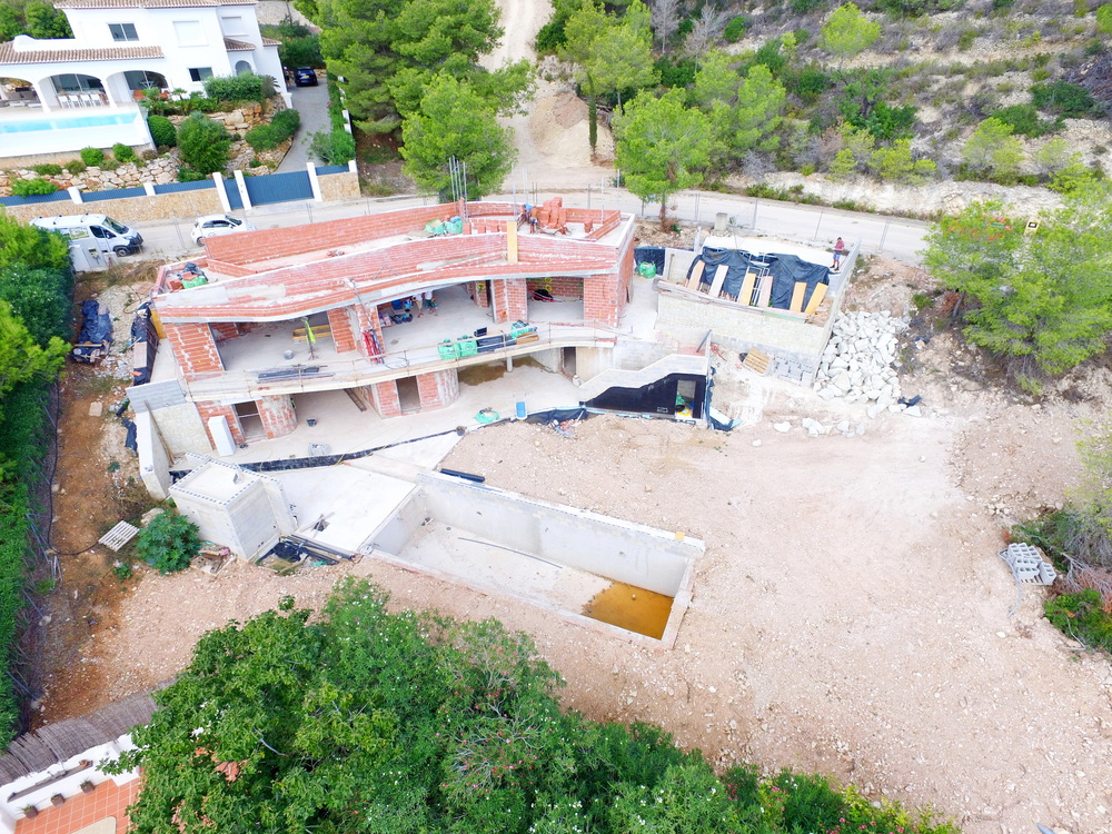 Aerial view of a multi-level villa under construction featuring red brick masonry, concrete foundations, and an excavated swimming pool area on a hillside.