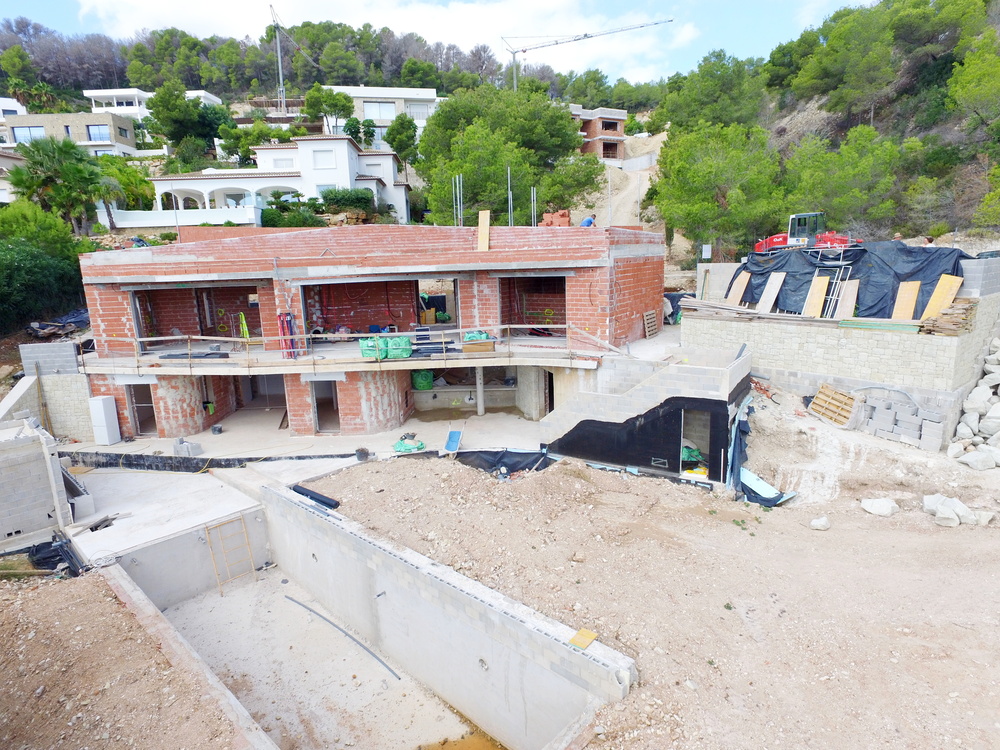 Construction phase of a two-story villa featuring a red brick structure and a concrete swimming pool shell on a terraced hillside plot surrounded by pine trees.