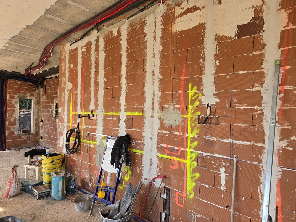 Construction phase showing a red clay brick wall with neon markings for electrical routing and exposed technical conduits on the ceiling.