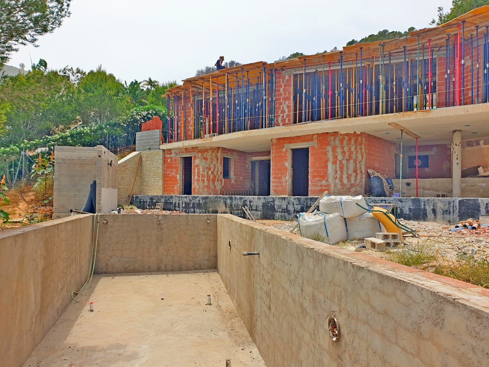 Construction site featuring a concrete swimming pool shell and a two-story brick building with metal shoring props and surrounding vegetation.