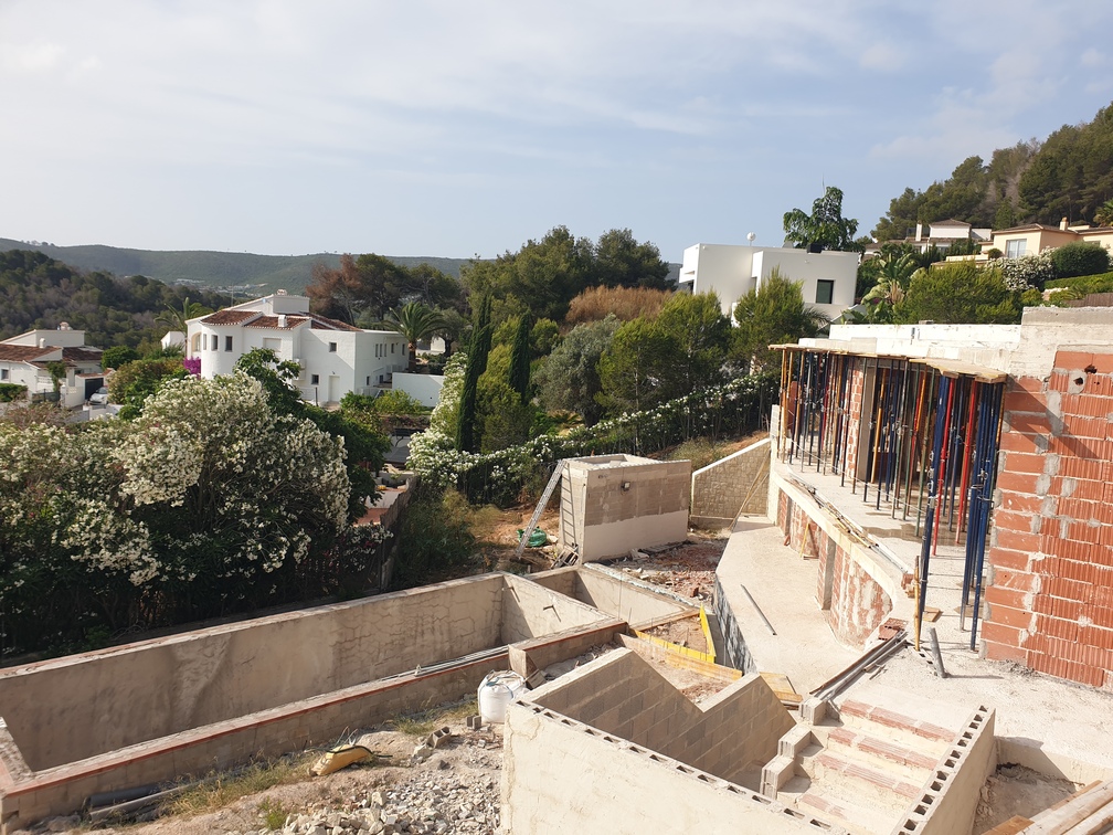 Construction site of a villa showing concrete foundations, brick walls with metal props, and a pool excavation overlooking a Mediterranean valley.