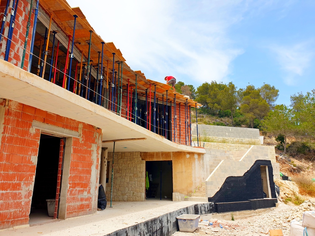 Modern villa under construction with red clay brick walls, concrete slabs, and metal shoring props. The site features a stone-clad wall and a concrete block staircase.