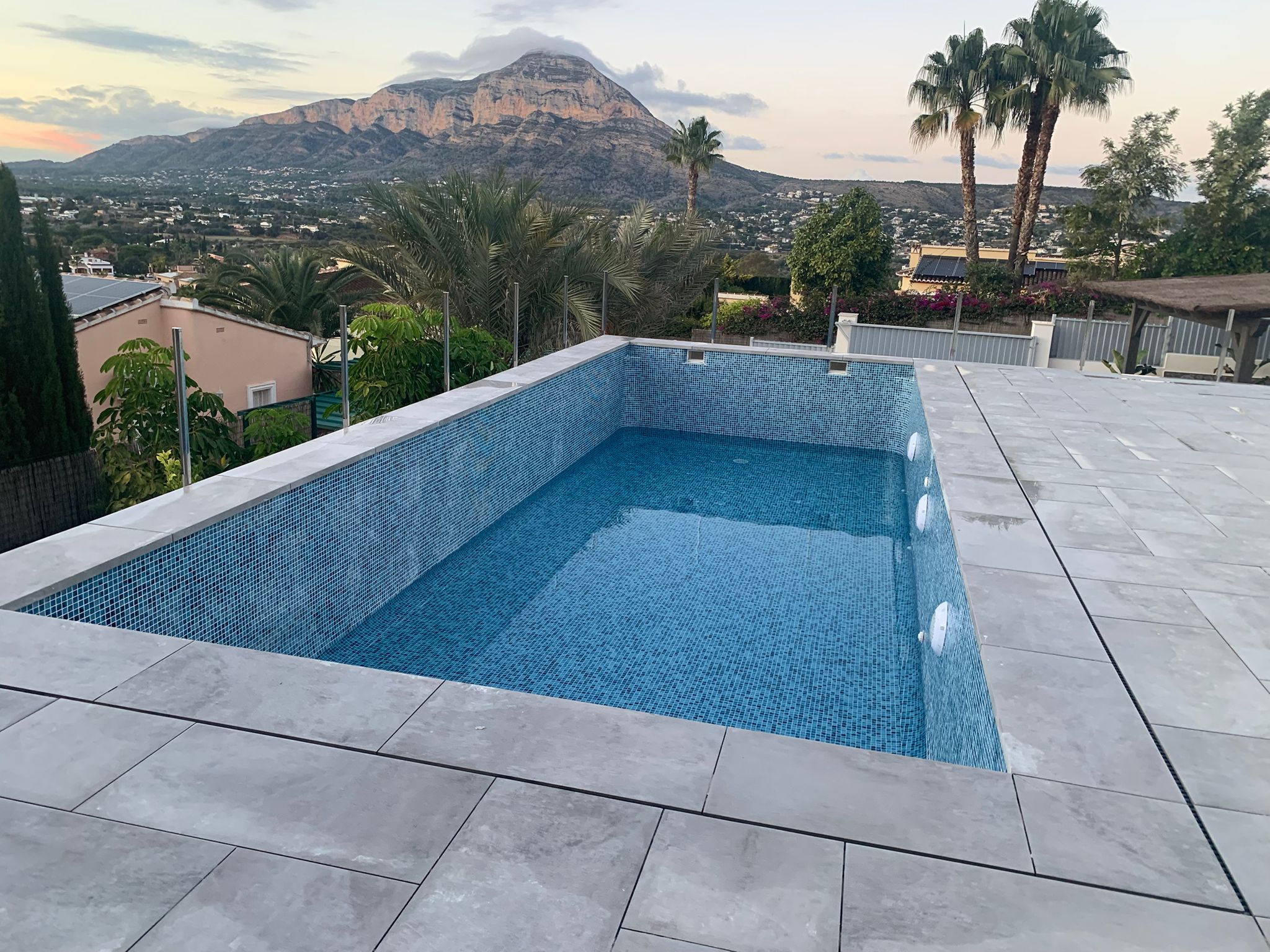 Elevated rectangular pool with blue mosaic tiles and grey stone terrace, featuring glass railings and panoramic views of the Montgó mountain.