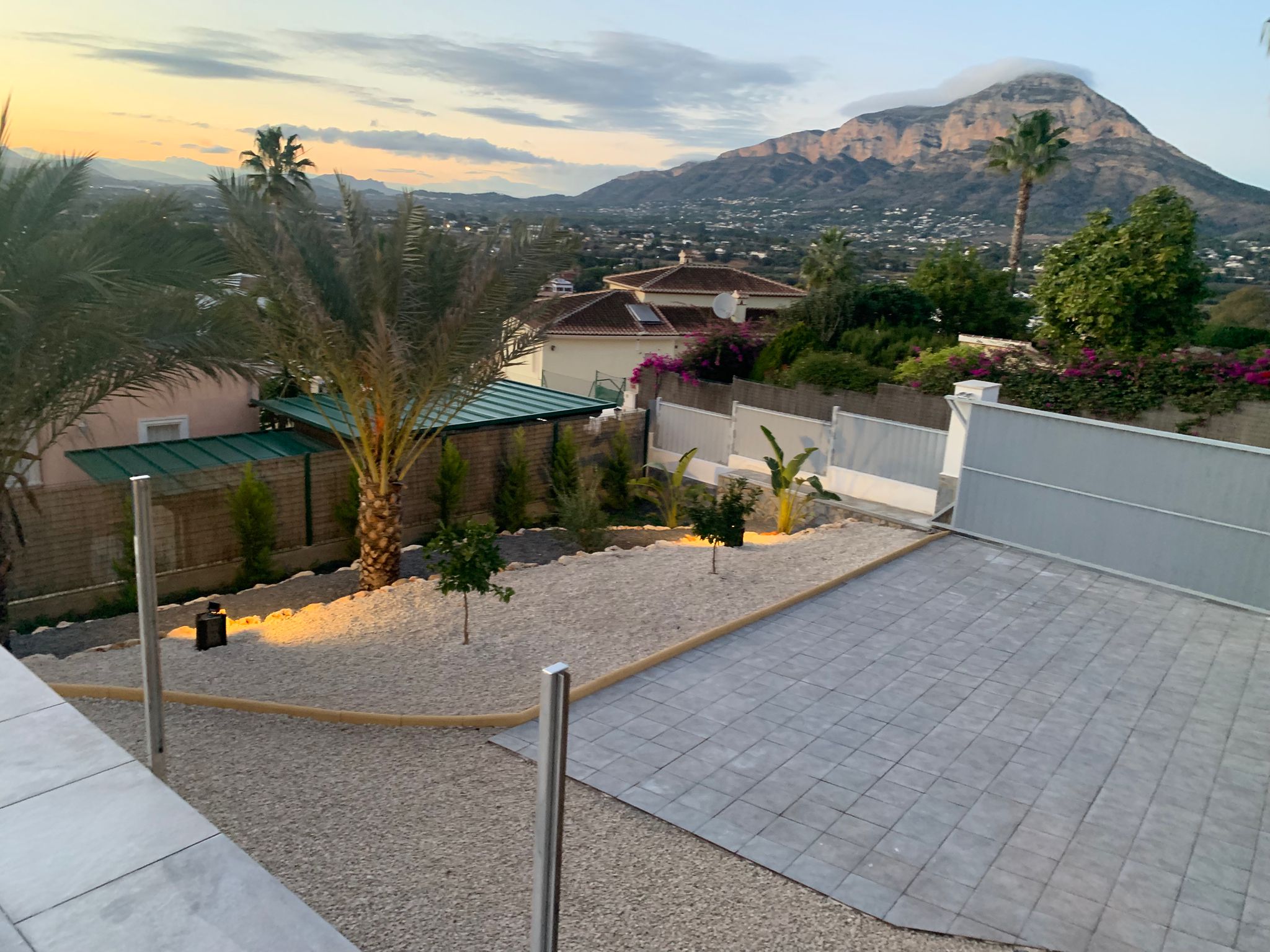 Elevated view of a landscaped garden with gravel, palm trees, and a paved driveway, featuring panoramic mountain views at sunset.