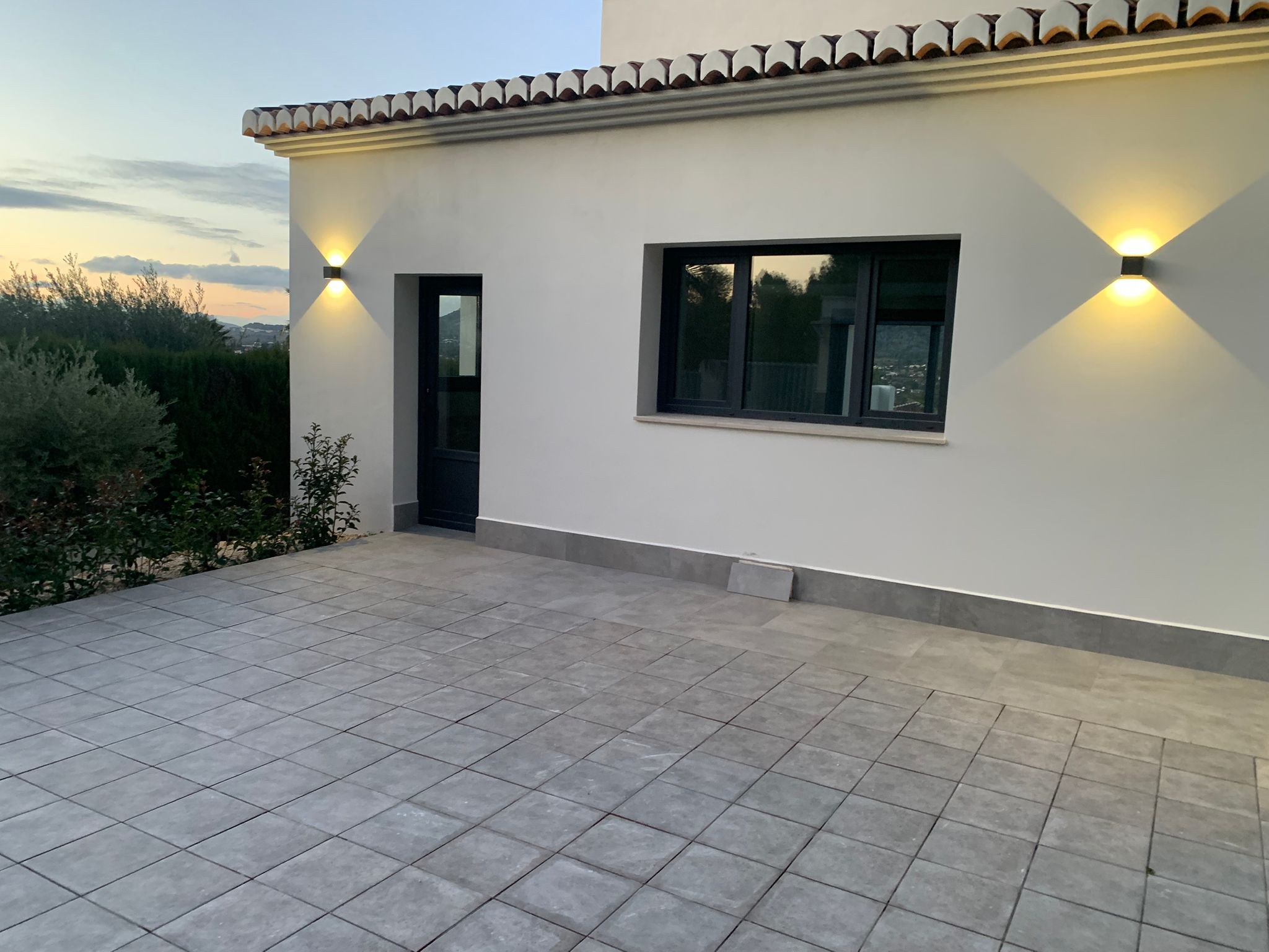 Exterior terrace with grey stone tiling, white facade, and modern wall lighting. Features dark-framed windows and a view of the surrounding hills at dusk.