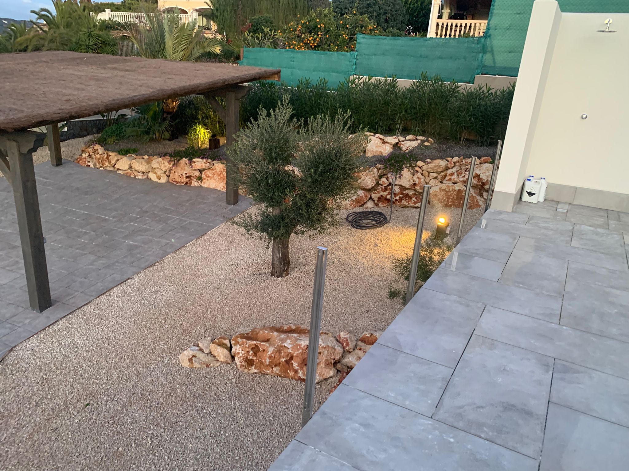 Mediterranean garden featuring a gravel area with an olive tree, natural stone borders, a thatched pergola, and grey stone tiling with an outdoor shower.