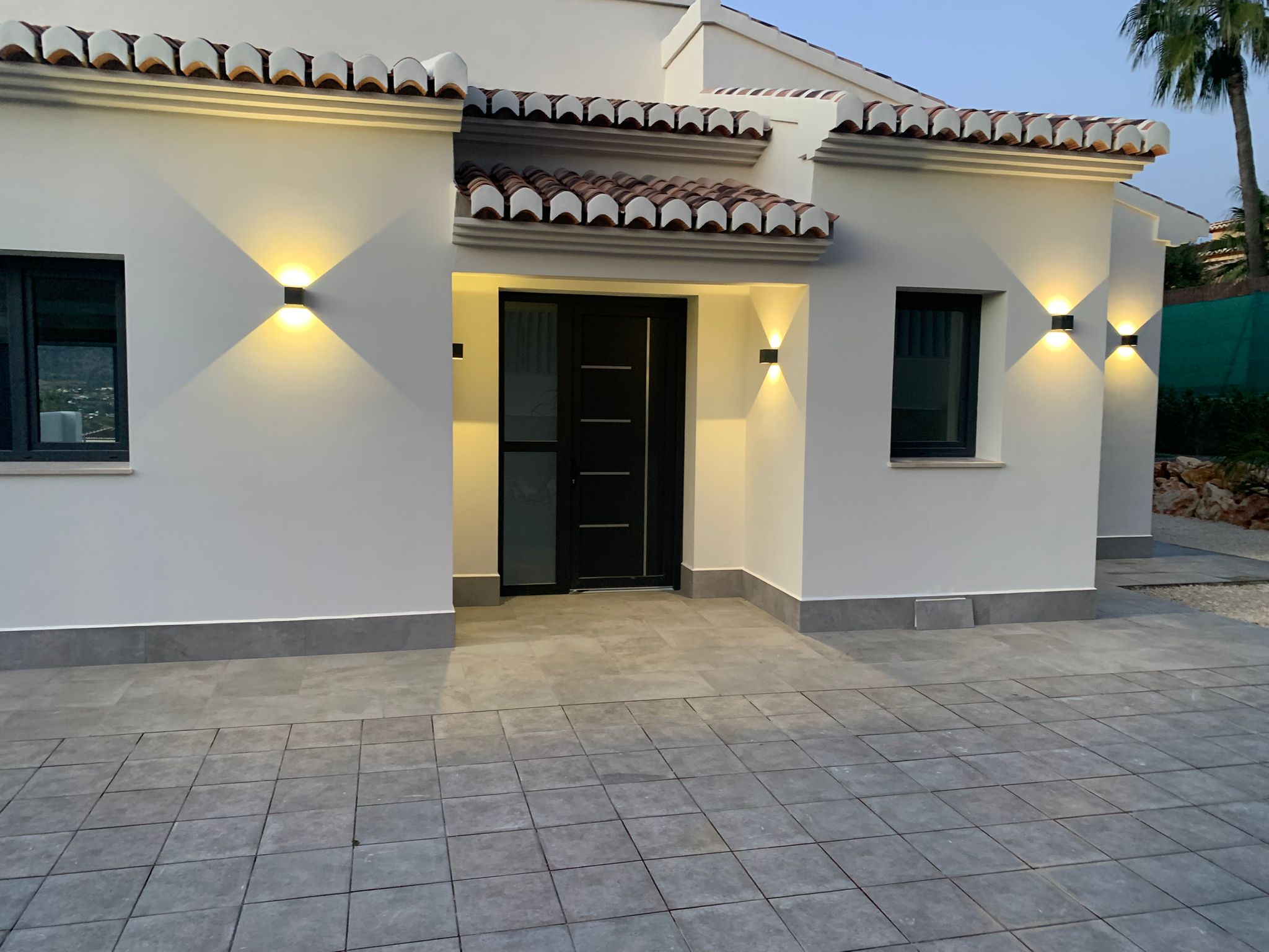 Modern villa entrance with a black door, white facade, and grey tiled patio. Symmetrical wall sconces provide warm lighting under the terracotta tile roof.