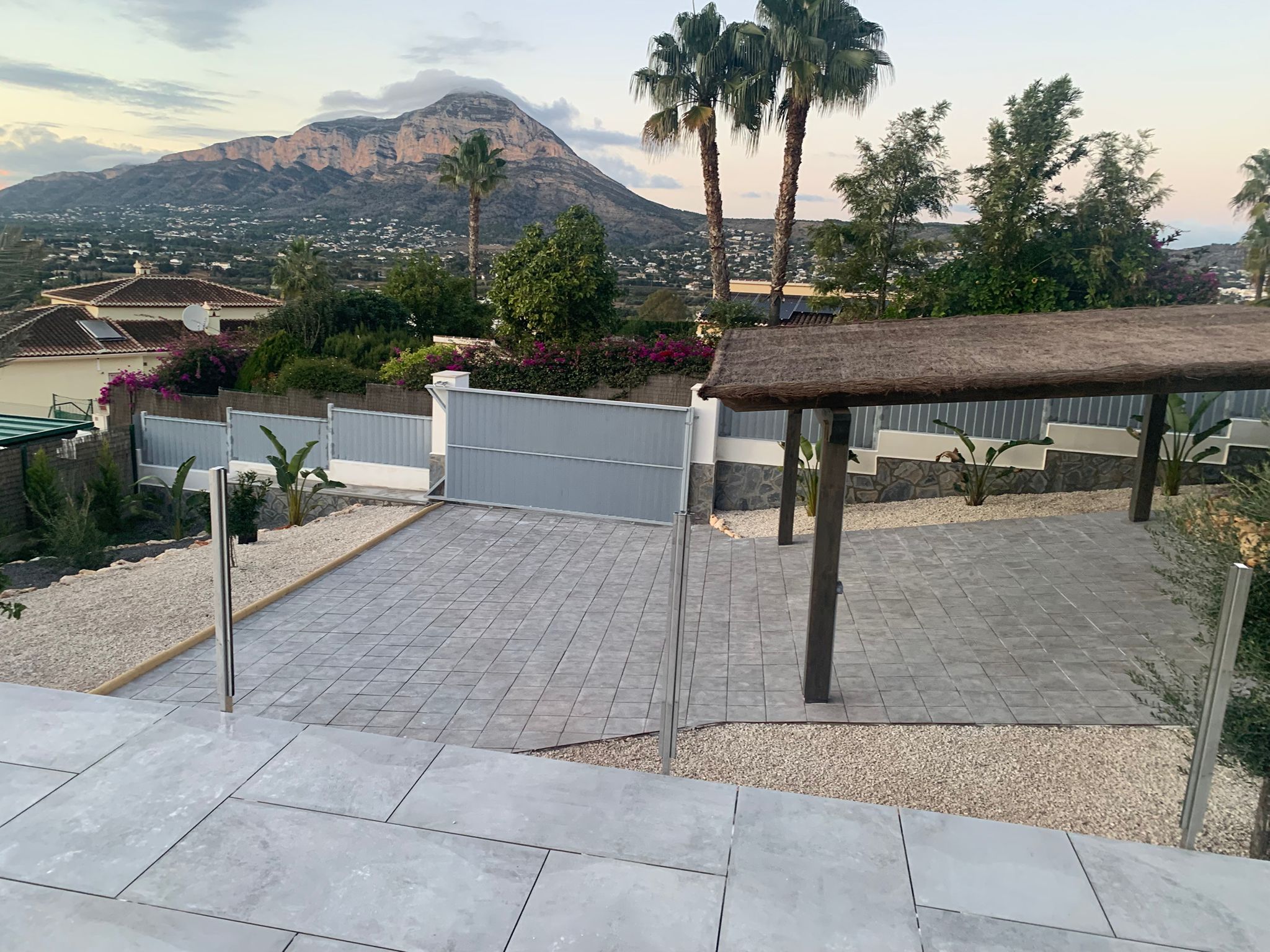 Paved outdoor driveway with grey tiling, a wooden pergola featuring a thatched roof, and panoramic views of the mountain range at dusk.
