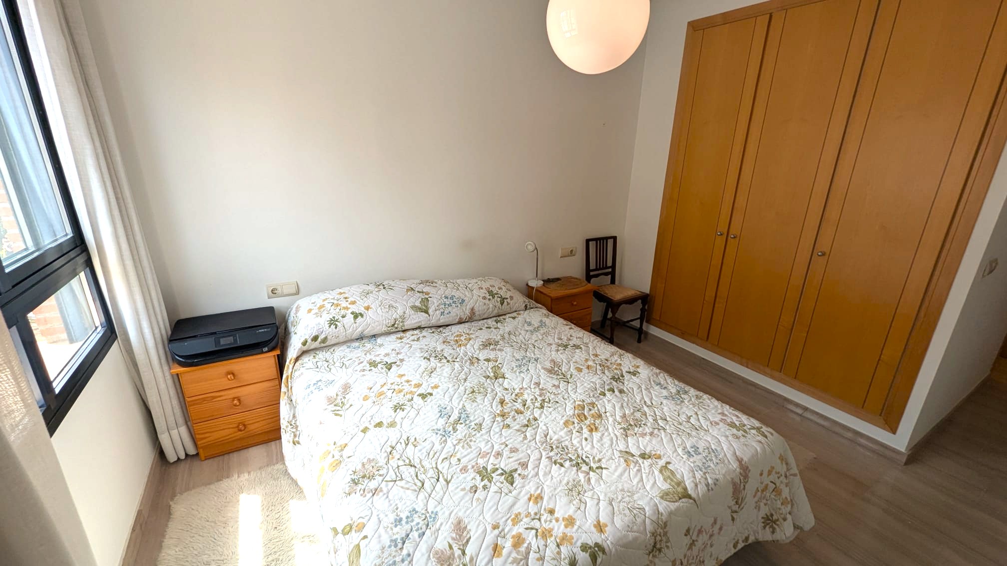 Bedroom featuring a built-in four-door wooden wardrobe, double bed, and matching nightstands. It includes light-colored wood flooring and a black-framed window.