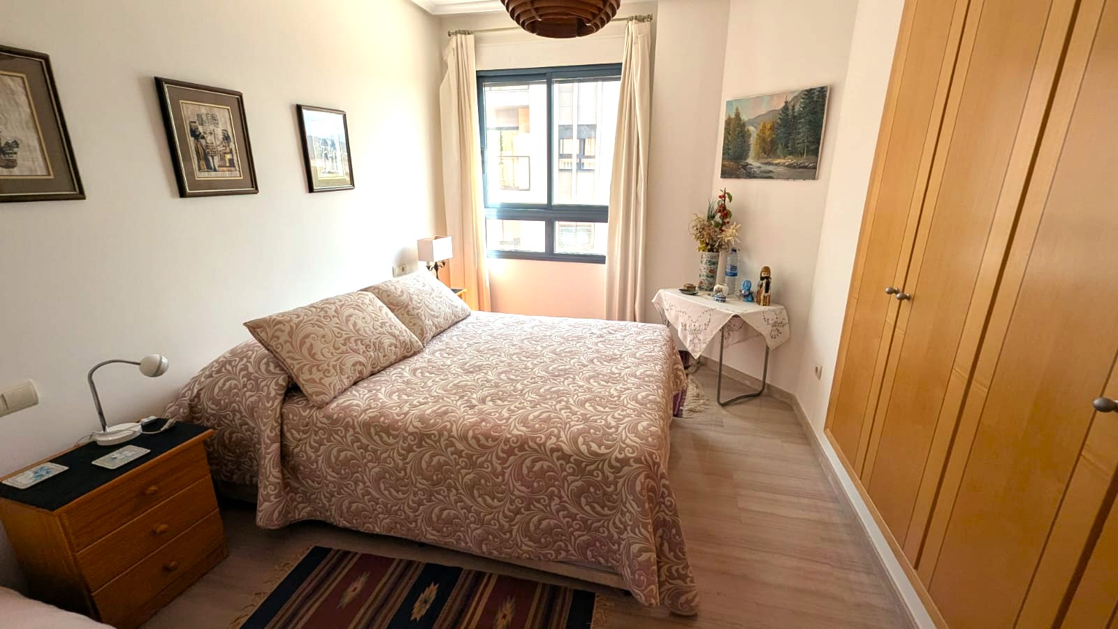 Bedroom featuring built-in wooden wardrobes, light laminate flooring, and a window for natural light. Includes functional furniture and classic decor in warm tones.