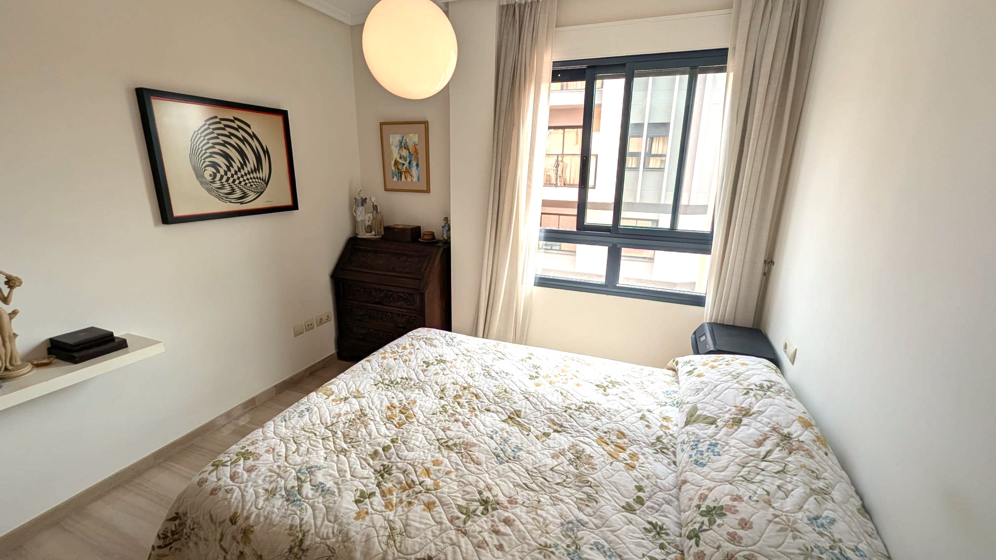 Bright bedroom featuring a dark wood antique bureau, a large window with neutral curtains, and a spherical pendant light over a floral-patterned bed.