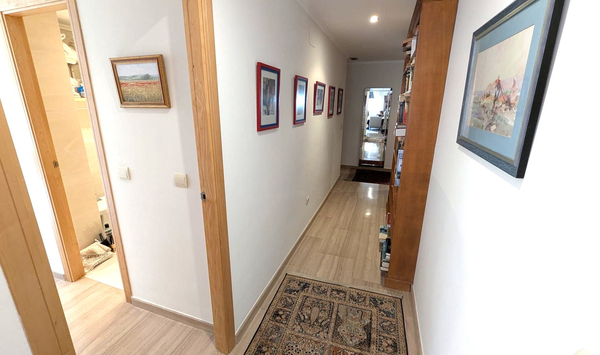Bright hallway featuring polished stone flooring, wooden door frames, and a built-in wooden bookshelf. Decorated with framed art and recessed lighting.