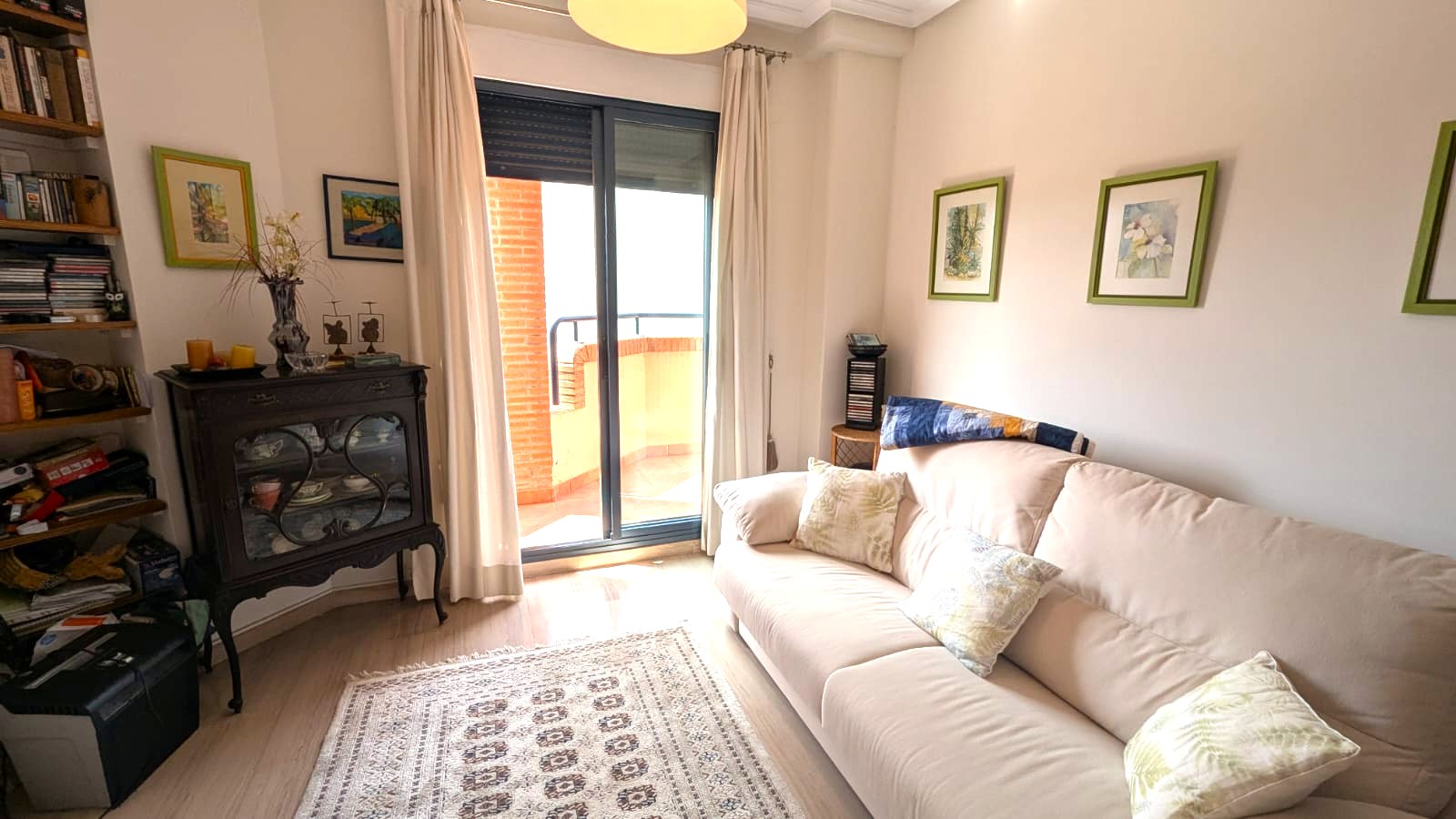 Bright living area featuring a beige sofa, vintage dark wood display cabinet, and sliding glass doors opening onto a brick-walled balcony.