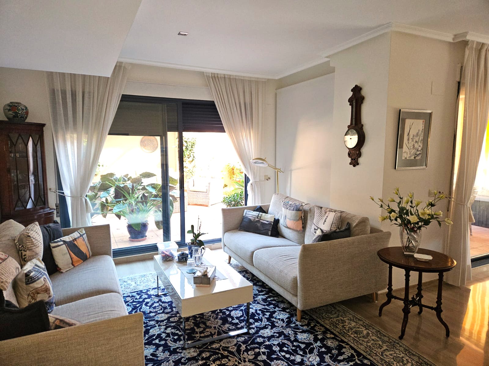 Bright living area with two neutral sofas, a patterned blue rug, and large sliding glass doors leading to a private terrace with lush greenery.
