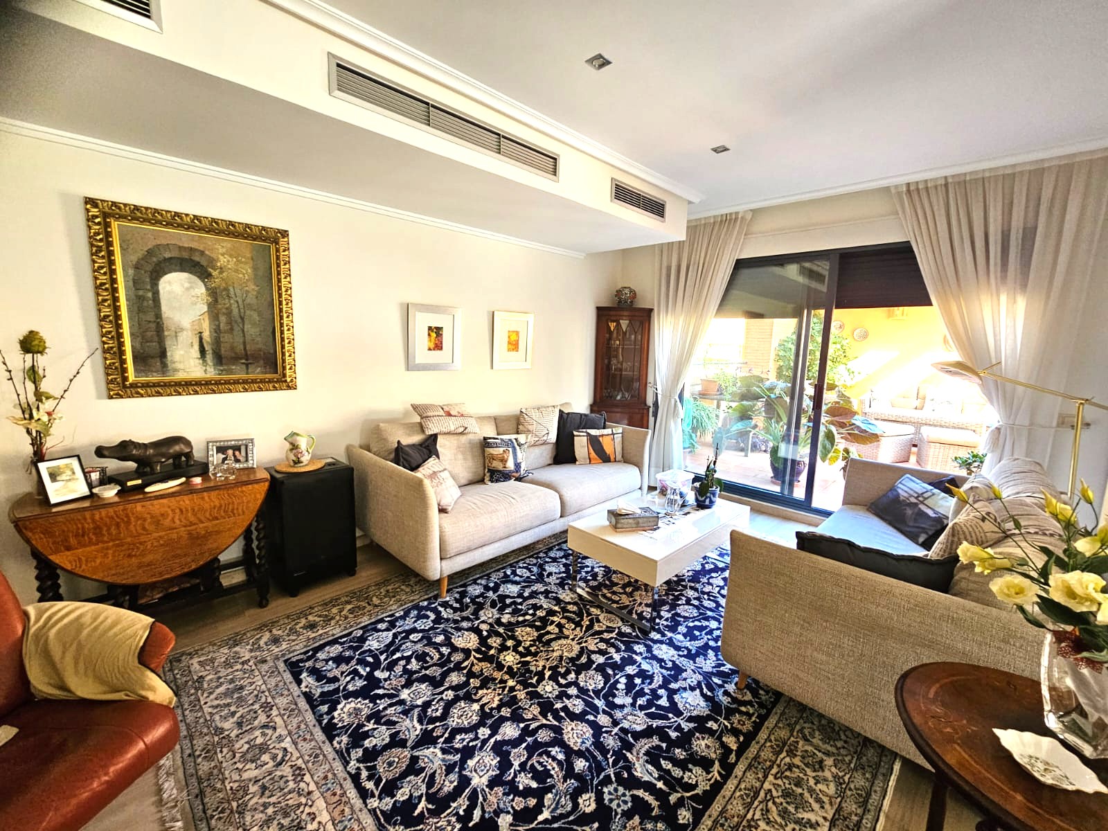 Bright living room featuring fabric sofas, a patterned blue rug, and direct terrace access. Equipped with integrated air conditioning vents and recessed ceiling lighting.