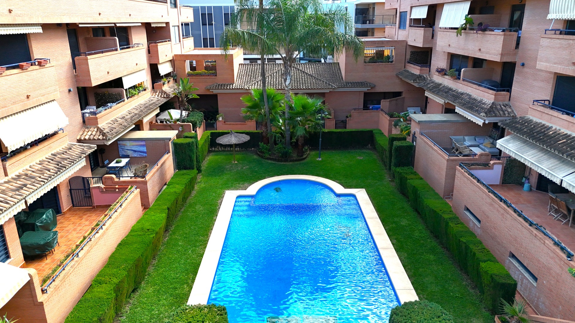 Central communal area featuring a rectangular swimming pool surrounded by manicured lawns, palm trees, and brick-clad apartment buildings.