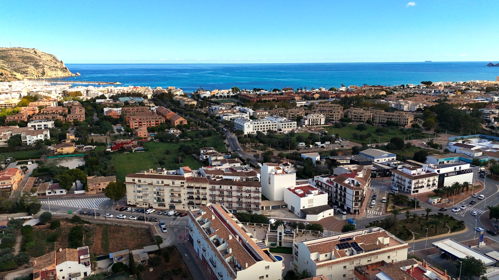 Elevated panoramic view of Jávea's urban landscape, featuring Mediterranean architecture, the coastal harbor area, and the blue sea under a clear sky.