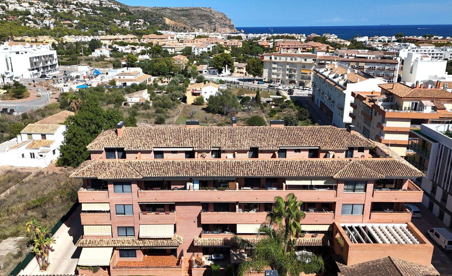 Elevated view of a brick apartment complex with terracotta roofing, featuring balconies and awnings, overlooking the town of Jávea and the Mediterranean Sea.